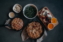 A dark tabletop features an assortment of dishes including a skillet of spaghetti topped with meat sauce and shredded cheese, a round loaf of sesame bread in a cast-iron pan, a bowl of green beans, marinated cheese cubes, grated parmesan, a dessert plate with berry-topped meringue or cheesecake, and two bowls of orange custard.
