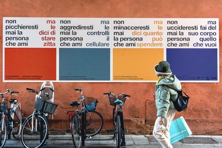 A street scene featuring a large colorful wall poster with text in Italian advocating against violence towards women. Several bicycles are parked against the wall, and a person in a green jacket and hat is walking by carrying a shopping bag.