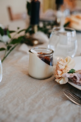 A small glass jar filled with a creamy dessert sits on a textured tablecloth, accompanied by a decorative flower and silverware. The background includes a few dark colored candles and some blurred greenery, contributing to an elegant and cozy dining atmosphere.