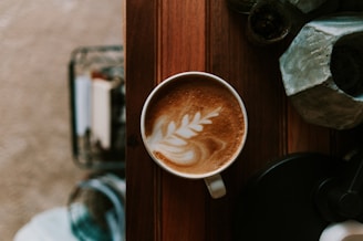 A vibrant coffee cup with latte art placed on a rustic wooden table, surrounded by fresh pastries and a small potted plant.