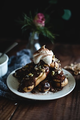 A plate of éclairs drizzled with rich chocolate sauce is garnished with chopped nuts and topped with a dollop of whipped cream. The setting is moody and rustic, with a dark background and a blurred flower arrangement adding a touch of elegance. The wooden table and subtle lighting create a cozy atmosphere.