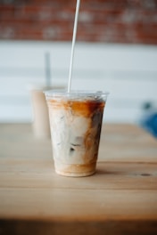 A clear plastic cup filled with iced coffee or a similar beverage, showing a gradient of dark brown, creamy white, and swirling textures. The cup is placed on a light wooden table, and a white straw is inserted. The background features a blurred portion of another drink and a white and brick wall.