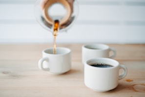 Three white ceramic mugs rest on a wooden table, with one being filled with coffee from a glass carafe. The background features a white, blurred setting, creating a minimalist and clean look.