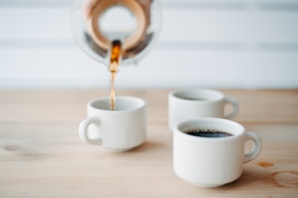 Three white ceramic mugs rest on a wooden table, with one being filled with coffee from a glass carafe. The background features a white, blurred setting, creating a minimalist and clean look.