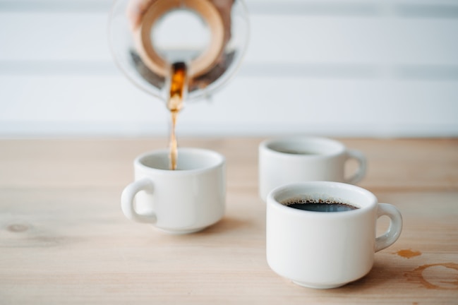 A close-up of a sleek, white minimalist mug filled with steaming coffee on a wooden table.