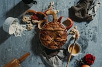 A freshly baked pie with a golden brown crust is surrounded by scattered apple peels, a small bowl of flour, a wooden rolling pin, a paintbrush, and a cup of caramel sauce. A red flower and a whole apple add a touch of color amidst the rustic setup. The textured gray surface and a piece of dark cloth create a homely, cozy atmosphere.