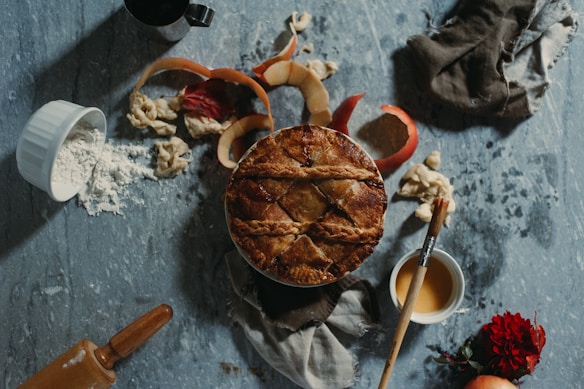 A freshly baked pie with a golden brown crust is surrounded by scattered apple peels, a small bowl of flour, a wooden rolling pin, a paintbrush, and a cup of caramel sauce. A red flower and a whole apple add a touch of color amidst the rustic setup. The textured gray surface and a piece of dark cloth create a homely, cozy atmosphere.
