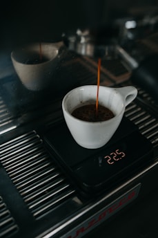 A white cup is placed on a digital scale as coffee is being brewed into it from an espresso machine. The digital display shows the weight measured in grams. The espresso machine's metal surface reflects light, indicating a clean, polished look.