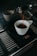 A white cup is placed on a digital scale as coffee is being brewed into it from an espresso machine. The digital display shows the weight measured in grams. The espresso machine's metal surface reflects light, indicating a clean, polished look.
