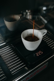 A white cup is placed on a digital scale as coffee is being brewed into it from an espresso machine. The digital display shows the weight measured in grams. The espresso machine's metal surface reflects light, indicating a clean, polished look.