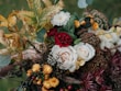 A detailed floral arrangement featuring a variety of flowers and foliage. Prominent elements include pale roses, deep red and yellow blossoms, surrounded by green leaves and a mix of dried and fresh elements. The overall composition appears lush and full, creating a harmonious blend of colors and textures.