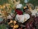 A detailed floral arrangement featuring a variety of flowers and foliage. Prominent elements include pale roses, deep red and yellow blossoms, surrounded by green leaves and a mix of dried and fresh elements. The overall composition appears lush and full, creating a harmonious blend of colors and textures.