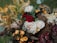 A detailed floral arrangement featuring a variety of flowers and foliage. Prominent elements include pale roses, deep red and yellow blossoms, surrounded by green leaves and a mix of dried and fresh elements. The overall composition appears lush and full, creating a harmonious blend of colors and textures.