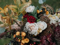 A detailed floral arrangement featuring a variety of flowers and foliage. Prominent elements include pale roses, deep red and yellow blossoms, surrounded by green leaves and a mix of dried and fresh elements. The overall composition appears lush and full, creating a harmonious blend of colors and textures.