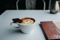 A white coffee cup filled with latte art featuring a heart design sits on a light table. A piece of chocolate rests on the edge of the cup. Next to the coffee, there is a brown chocolate bar wrapper with a modern design including the words 'Madagascar' and '75% cacao'. A metallic vase is visible in the background.