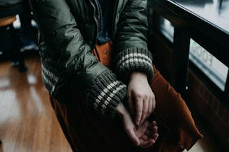 A person is sitting with hands folded on their lap, wearing a green jacket with plaid cuffs and rust-colored pants. The setting appears to be indoors, near a window with wooden flooring visible.