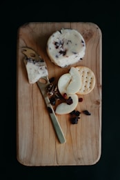 A rustic wooden cutting board displays a round wheel of cheese, a slice of which is cut and rests on a cheese knife. Nearby, there are sliced pear pieces, a round cracker, and scattered dried fruits.
