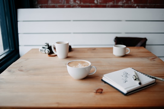 A wooden table with three cups of coffee, one in the foreground featuring latte art. A notebook with a pen rests on the table, and there is a vintage camera in the background. The setting has a cozy, rustic atmosphere.