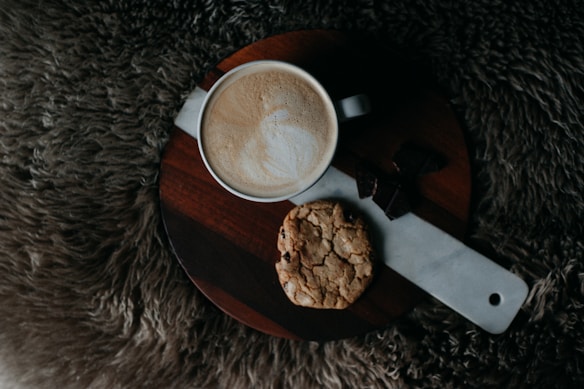 A cup of coffee with a frothy top placed on a wooden tray alongside a chocolate chip cookie. Dark chocolate pieces are also present on the tray. The background features a textured, fur-like surface.