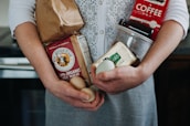 A person is holding various groceries, including a paper bag, organic whole wheat flour, eggs, a carton of Kerrygold butter, and a red coffee package. The individual wears a white knitted cardigan over a grey dress.