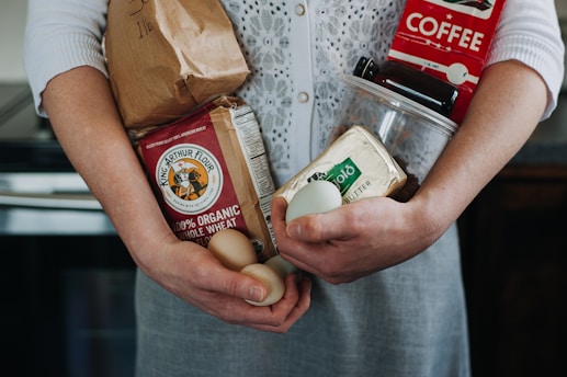 A person is holding various groceries, including a paper bag, organic whole wheat flour, eggs, a carton of Kerrygold butter, and a red coffee package. The individual wears a white knitted cardigan over a grey dress.