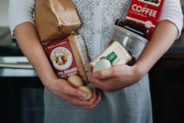 A person is holding various groceries, including a paper bag, organic whole wheat flour, eggs, a carton of Kerrygold butter, and a red coffee package. The individual wears a white knitted cardigan over a grey dress.
