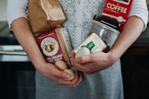 A person is holding various groceries, including a paper bag, organic whole wheat flour, eggs, a carton of Kerrygold butter, and a red coffee package. The individual wears a white knitted cardigan over a grey dress.