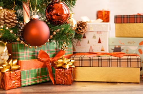 A festive arrangement of wrapped Christmas gifts in assorted patterns and colors sits beneath a section of a decorated Christmas tree. The tree is adorned with shiny red baubles, gold pine cones, and other ornaments. The presents feature various wrapping paper designs, including plaid and holiday-themed patterns, topped with shiny ribbons and bows.