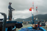 Two people are working on a fishing boat near the coast, with one person handling fishing lines and the other securing items on the boat. The boat is vibrantly painted blue and adorned with flags, including a prominent red flag with a yellow star. The scene is set against a cloudy sky and mountainous backdrop.