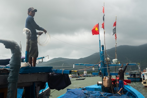 Two people are working on a fishing boat near the coast, with one person handling fishing lines and the other securing items on the boat. The boat is vibrantly painted blue and adorned with flags, including a prominent red flag with a yellow star. The scene is set against a cloudy sky and mountainous backdrop.