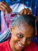 A person with braided hair smiles warmly while another pair of hands is styling their hair. The braids are intricate and feature a mix of black and blue tones. The person wears a red shirt and appears cheerful, while the background is slightly blurred with hints of colorful patterns.