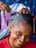 A person with braided hair smiles warmly while another pair of hands is styling their hair. The braids are intricate and feature a mix of black and blue tones. The person wears a red shirt and appears cheerful, while the background is slightly blurred with hints of colorful patterns.