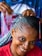A person with braided hair smiles warmly while another pair of hands is styling their hair. The braids are intricate and feature a mix of black and blue tones. The person wears a red shirt and appears cheerful, while the background is slightly blurred with hints of colorful patterns.