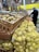 Wooden crates and yellow mesh bags filled with potatoes are neatly arranged on a grocery store display. In the background, a person wearing a hairnet is selecting produce from an adjacent section, suggesting a shopping environment.