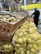 Wooden crates and yellow mesh bags filled with potatoes are neatly arranged on a grocery store display. In the background, a person wearing a hairnet is selecting produce from an adjacent section, suggesting a shopping environment.