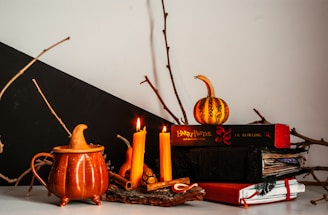 A cozy autumnal scene features a small pumpkin-shaped teapot, three lit orange candles, a few twigs, and a small decorative pumpkin. There are several stacked books, including a notable Harry Potter title by J.K. Rowling, creating a warm and inviting atmosphere.