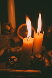 Two lit candles are positioned in the foreground, surrounded by decorative elements such as pine cones and a dried citrus slice. The candles emit a soft, warm glow, casting flickering shadows and creating an intimate atmosphere.