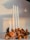 A decorative arrangement featuring four white candles of varying heights, one of which is lit. The candles are set on a piece of driftwood, adorned with dried orange slices, pine cones, and gold-painted ornaments. The setup exudes a warm and rustic ambiance.