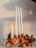 A decorative arrangement featuring four white candles of varying heights, one of which is lit. The candles are set on a piece of driftwood, adorned with dried orange slices, pine cones, and gold-painted ornaments. The setup exudes a warm and rustic ambiance.