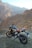A motorcycle is parked on a rocky terrain with a backdrop of rugged mountains. The bike has a sleek design with a combination of black and gold colors, and it stands on a dirt path with an open road visible. The setting creates a sense of adventure and exploration.
