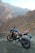 A motorcycle is parked on a rocky terrain with a backdrop of rugged mountains. The bike has a sleek design with a combination of black and gold colors, and it stands on a dirt path with an open road visible. The setting creates a sense of adventure and exploration.