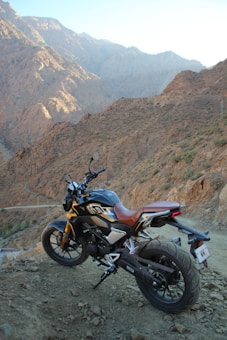 A motorcycle is parked on a rocky terrain with a backdrop of rugged mountains. The bike has a sleek design with a combination of black and gold colors, and it stands on a dirt path with an open road visible. The setting creates a sense of adventure and exploration.