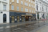 A historic hotel located on a city street, featuring a beige facade with multiple windows. The entrance is marked with a sign reading 'Hotel Stefanie' above a glass door. Adjacent buildings have ornate architecture, and tram tracks run along the wet pavement in front.