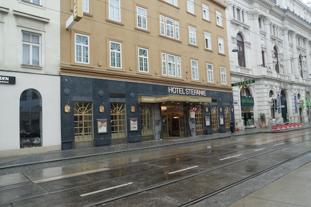A historic hotel located on a city street, featuring a beige facade with multiple windows. The entrance is marked with a sign reading 'Hotel Stefanie' above a glass door. Adjacent buildings have ornate architecture, and tram tracks run along the wet pavement in front.