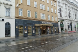 A historic hotel located on a city street, featuring a beige facade with multiple windows. The entrance is marked with a sign reading 'Hotel Stefanie' above a glass door. Adjacent buildings have ornate architecture, and tram tracks run along the wet pavement in front.