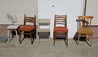An arrangement of different chairs and a small table against a wall. The two wooden chairs feature orange cushions, and another chair to the right has a plain wooden seat. A small square table on the left has a simple design and white top. The background appears to be a white door set into a beige wall.