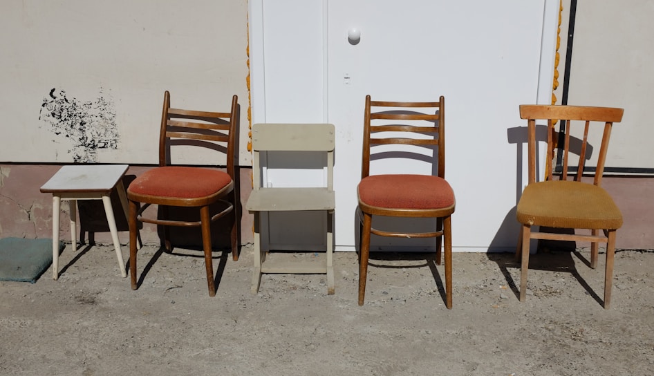 An arrangement of different chairs and a small table against a wall. The two wooden chairs feature orange cushions, and another chair to the right has a plain wooden seat. A small square table on the left has a simple design and white top. The background appears to be a white door set into a beige wall.