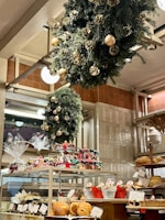 A bakery display filled with various pastries and baked goods, decorated with festive wreaths adorned with gold ornaments and pinecones. The shelves hold an assortment of bread and wrapped gift baskets, while a carousel centerpiece adds a whimsical touch.