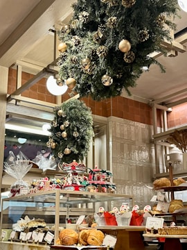 A bakery display filled with various pastries and baked goods, decorated with festive wreaths adorned with gold ornaments and pinecones. The shelves hold an assortment of bread and wrapped gift baskets, while a carousel centerpiece adds a whimsical touch.