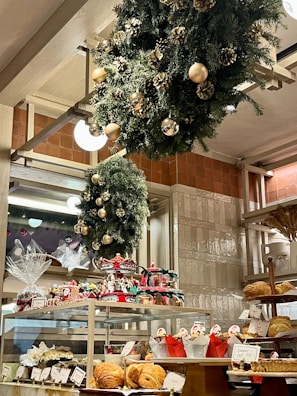A bakery display filled with various pastries and baked goods, decorated with festive wreaths adorned with gold ornaments and pinecones. The shelves hold an assortment of bread and wrapped gift baskets, while a carousel centerpiece adds a whimsical touch.